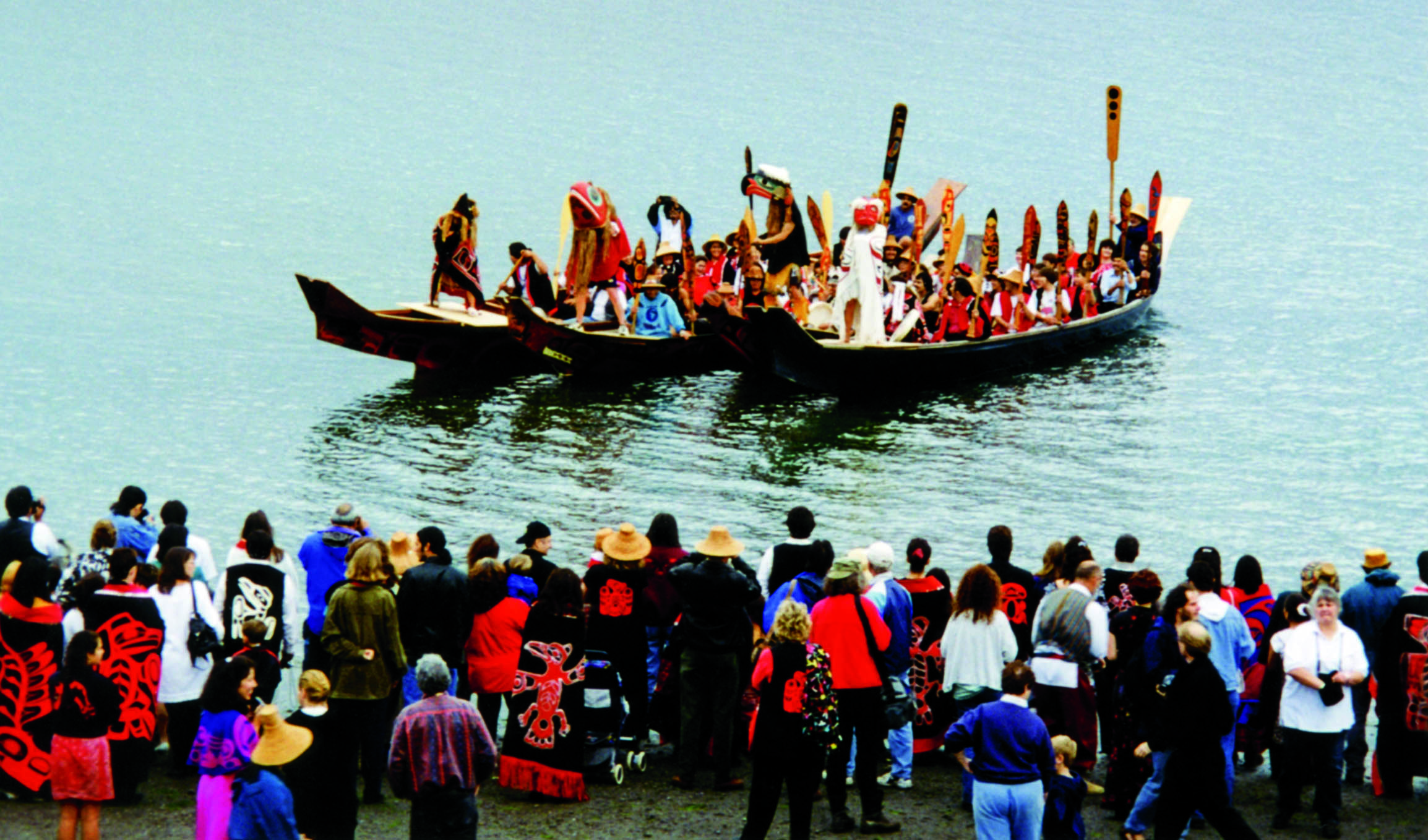 Two canoes filled with paddlers arrive at shore to a throng of people watching. Paddles are pointing up and dancers in regalia and masks stand on platforms built on the canoes' prow.