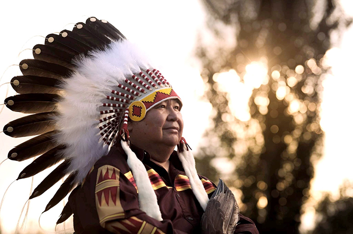 A man stands with his arms crossed across his chest. He has an eagle wing in his hand. He wears a shirt with geometric designs acress the front near his shoulder and down his arms. On his head is a feathered headdress. The same designs as on his shirt is beaded on the headband of the headdress.