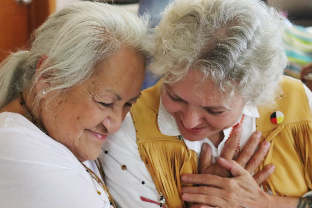 Two older woman share a time together. One has brought the others hand and placed it over her heart. They bring their heads together with affection.