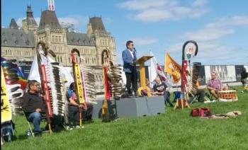 Eagle staffs are lined up on the lawn in front of Parliament Hill. The Parliament building can be seen in the backgroup. A man stands at a podium to speak. 