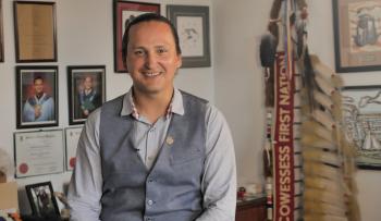 A smiling man wearing a shirt with a suit vest over top sits perched on the edge of a desk. On the walls behind are photographs of graduates and diplomas. Behind his is the eagle staff of the Cowesses First Nations.