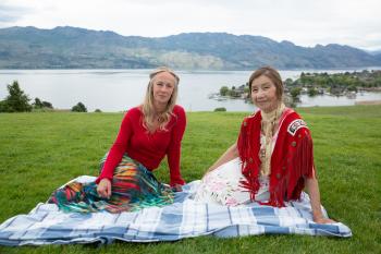 Two women sit on a blue checkered blanket on a green grassy area in front of a lake.