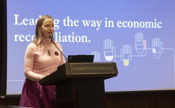 A woman stands at a podium in front of a screen with the words "Leading the way in economic reconciliation" printed on it.