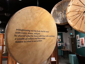Indigenous hide hand drums hang in a grouping and there are verses of a poem printed on each of them.