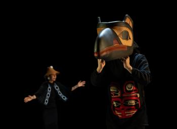 A dark stage is the backdrop for two dancers. A woman is seen in the background. And in front is a masked dancer. The mask is of a raven, and it is a huge piece the dancer has over his head.