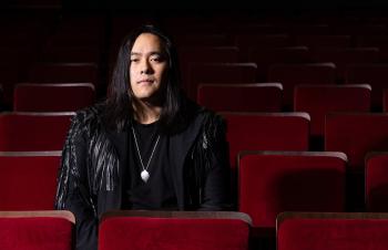 A man with long black hair sits alone in a theatre with red velvet chairs.