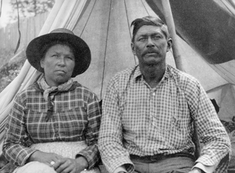A black and white photo of a woman and man sitting in front of a canvas tent.