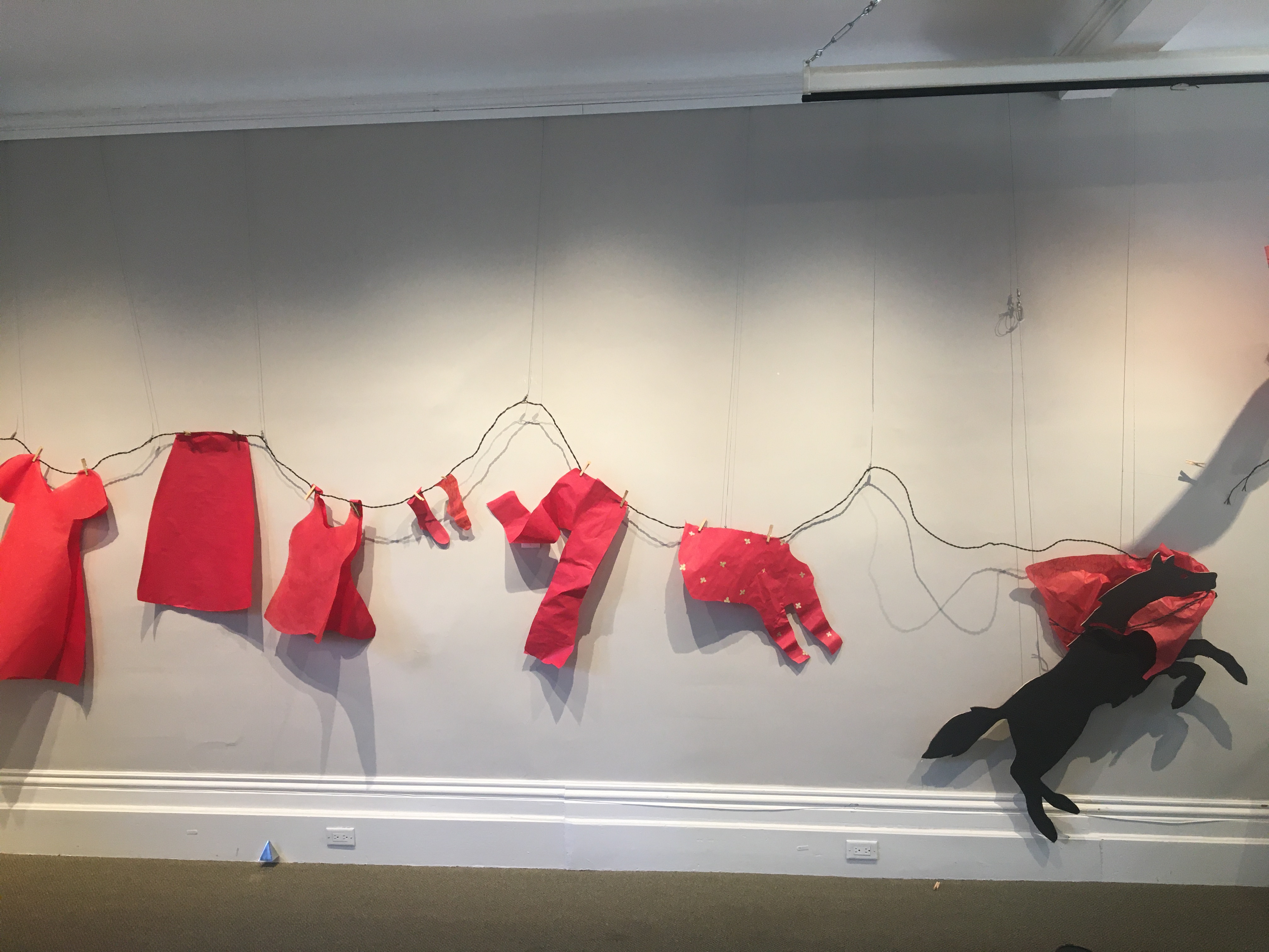 A wire serves as a clothes line with red paper cut-outs of pants, shirts and dresses hang along it.  At the end is a black silhouette of a wolf lunging with a red dress in its mouth.
