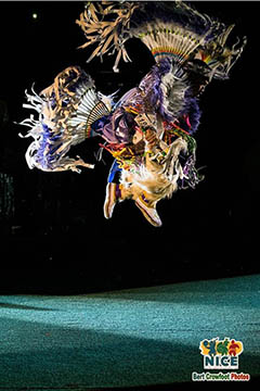 A fancy dancer is photographed in the air in a spotlight against a black background