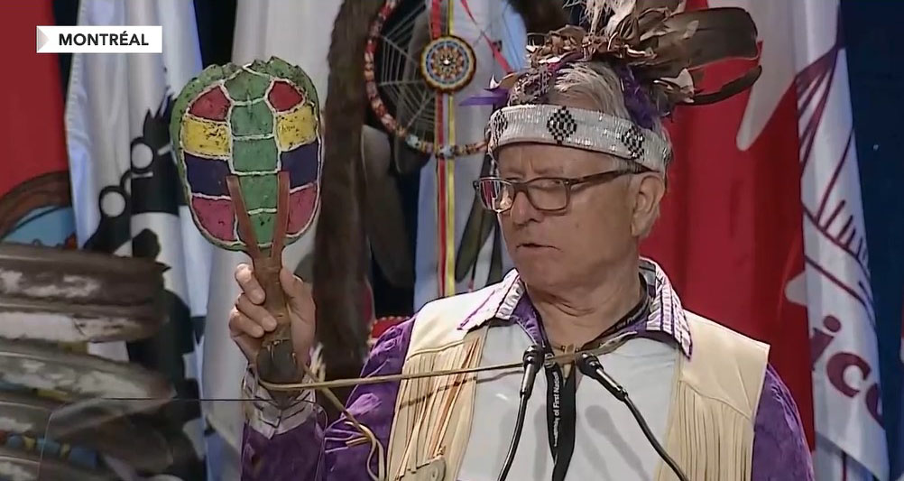A man in a beaded headdress with feathers holds up a painted turtle ratte as he speaks at a microphone.