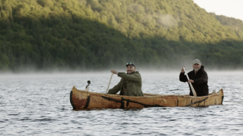 Two First Nations men paddle a birchbark canoe.