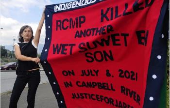 Mother of slain man holds up red button blanket with words on it calling out the RCMP for his death.