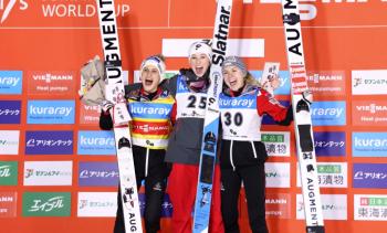 Winners of the women's World Cup ski jumping competition stand in front of an orange wall festooned with multi-coloured plaques with Japanese writing on them. The women hold their skis, bouquets and have their arms around each other. They smile wide with excitement.