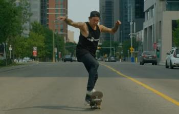 An Indigenous man in black rides a skateboard down a city street.