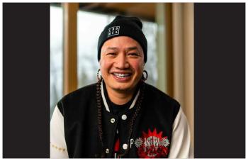 Patrick Mitsuing smiles into the camera. He's wearing a black toque that reads Land Back on it, and a black jacket with white sleeves. It has a logo that reads Powwow.