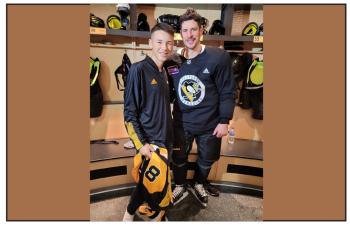 A teenager male stands next to an NHL hockey player in a Pittsburgh Penguins' uniform inside a dressing room. The teen holds a jersey with the number 18 on it.