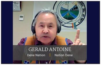A man is on a livestream call. He wears a buckskin beaded vest and earphones and holds his finger up as he speaks. Behind him is a flag of the Dene Nation. The logo has two rivers. Trees at the bottom, grasslands on eachside and mountains behind. It says "Land of the People."