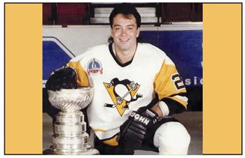 A man kneels beside a trophy, a big silver cup. The man wears a hockey jersey that is white with yellow sleeves. On the front is a logo of a penguin on skates with a hockey stick.