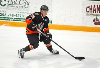 A hockey playing wearing a black uniform with orange accents has the puck on his stick.