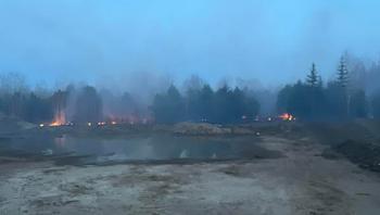 A wildfire can be seen flaming amongst the trees in the background while the earth has been scrapped of brush in the foreground as a pool of water remains on the bare ground.