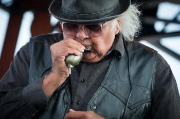 A man with grey hair bursting from beneath a pork pie hat plays harmonica into a mic. He wears all black and sunglasses.