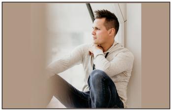 A man sits near a window and stares out. He leans on the wall behind him.