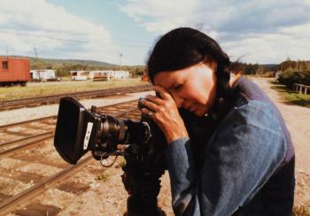 A woman sporting a long black braid that hangs over her shoulder stares into a film camera.