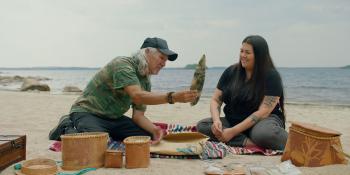 Two people sit on a beach with a body of water behind them. They are surrounded by a display of birchbark baskets, and the older man is holding some artifact for the younger woman to look at.
