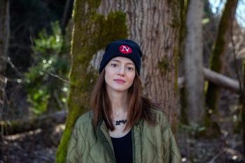 A young woman stands against the backdrop of old growth trees. Moss is growing up one side of a big tree directly behind her. She wears a black toque with an N in white on a red circle on the hat.