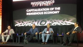 Four men sit on a stage in front of a wide screen on which is printed Capitalization of the Indigenous Economy over some art work in the West Coast style.