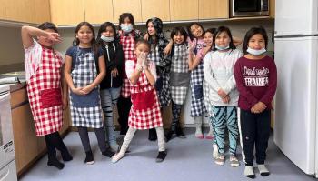 A group of children, mostly girls, dress in red or blue checked aprons in a bright kitchen.