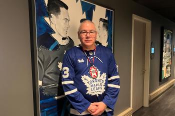 A man in a Toronto Maple Leafs jersey stands beside a large drawing featuring former Toronto Maple Leafs captain George Armstrong, whose mother was from the Kitigan Zibi Nation.