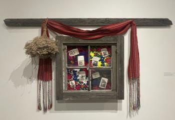 An old house board and window hangs on a gallery wall. Inside the frames of the window are photographs. A Métis sash is draped above the window an a bouquet of dried flowers hang upside down on the left side of the window frame.