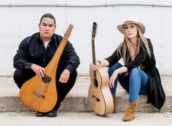 A man and a woman sit on a cement step in front of a white painted wall. Each hold a guitar. The man's guitar is a lap steel with a blunted headstock and a body that is round at the bottom but tapers up the neck.