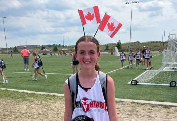A young tween girl smiles at the camera. She is wearing an Ontario jersey and a headband with two Canadian flags standing upright on it.