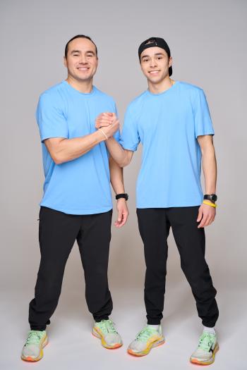 A man and his teen son clasp hand and smile towards the camera. They are in matching light blue t-shirts and the teen wears his ball cap backwards on his head.