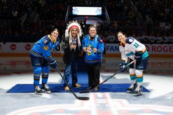 A man wearing a feather bonnet and a woman in a hockey jersey stand waiting to drop th puck for two women hockey players.