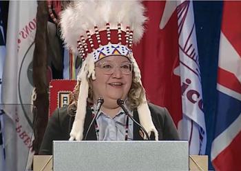 A woman in a feather bonnet stands at a podium tospeak.