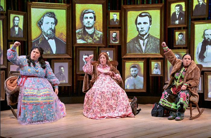 Three women in 1800s garb sit in chairs on a stage in front of framed portraits of men from the time period. The women have their right arms raised above their heads and hands making fists.