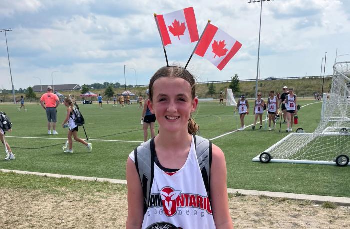 A young tween girl smiles at the camera. She is wearing an Ontario jersey and a headband with two Canadian flags standing upright on it.
