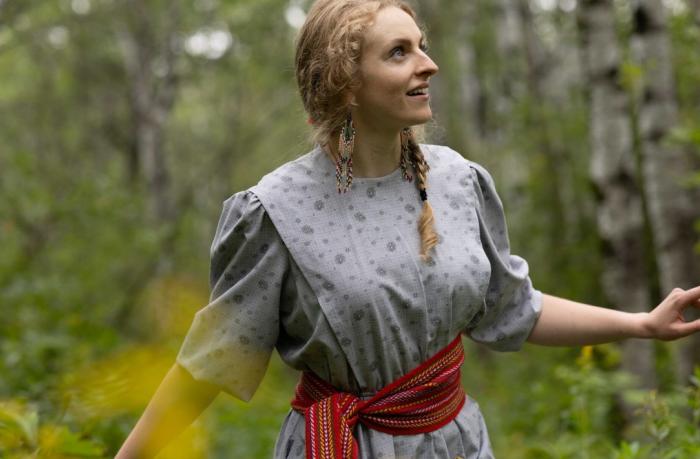 A young girl in a cotton dress with a Metis sash around her waist walks through the woods.