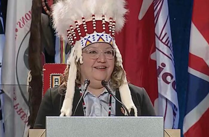 A woman in a feather bonnet stands at a podium tospeak.