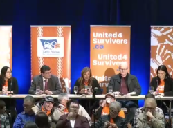 Five people sit at a long table in front of vertical orange panels with writing on them, including United4Survivors.ca and handprints with that same website printed on them.