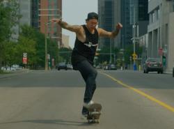 An Indigenous man in black rides a skateboard down a city street.