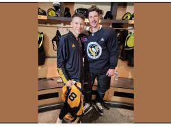 A teenager male stands next to an NHL hockey player in a Pittsburgh Penguins' uniform inside a dressing room. The teen holds a jersey with the number 18 on it.