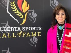 A woman stands against a grey wall with logos printed on it and the words Alberta Sports Hall of Fame. She wears a bright pink jacket and has a folded red blanket draped over her shoulder.