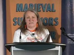 A woman stands at the podium with a panel behind her that reads Marieval Indian Residential School