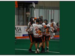 A jubilant group of lacrosse players meet and hug in a box arena.