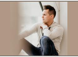 A man sits near a window and stares out. He leans on the wall behind him.