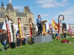 Eagle staffs are lined up on the lawn in front of Parliament Hill. The Parliament building can be seen in the backgroup. A man stands at a podium to speak. 